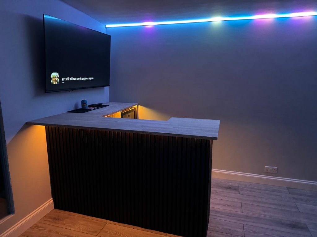Modern home bar with wood slat front panel, under-cabinet lighting, LED ceiling strip lights, and wall-mounted TV in a stylish entertainment room