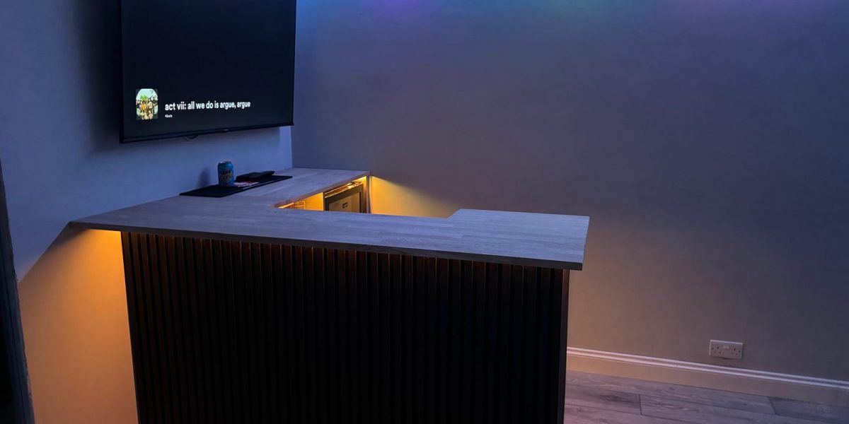 Modern home bar with wood slat front panel, under-cabinet lighting, LED ceiling strip lights, and wall-mounted TV in a stylish entertainment room